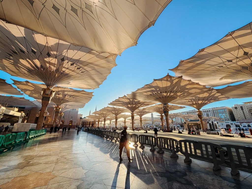 Sunlit view of architectural umbrellas in Medina with people walking, creating dramatic shadows.